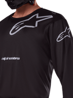 Alpinestars 2025 Racer Graphite Jersey, maglia da motocross nero e grigio, primo piano del pilota con marchio Alpinestars e loghi A-star grigi, materiale in politessuto tecnico per guida fuoristrada