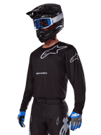 Pilota di motocross con Set Gear Alpinestars Racer Graphite 2025, maglia nera con loghi Alpinestars grigi, pantaloni neri coordinati con dettagli grigi, casco nero con occhiali blu e bianchi, guanti blu e grigi, abbigliamento tecnico per corse fuoristrada