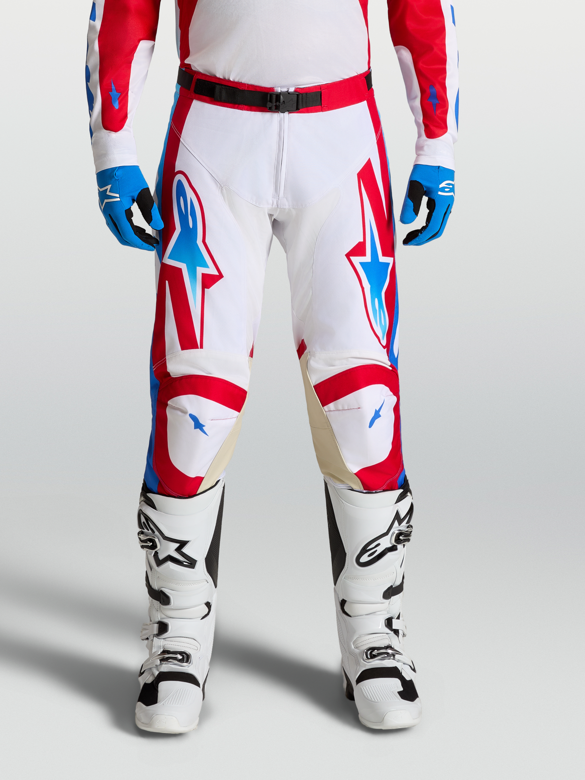 Alpinestars Pantaloni Racer Hunt, pantaloni motocross da uomo, bianco, rosso e blu, con gambe pre-curvate ergonomiche e pannelli in pelle sulle ginocchia per fuoristrada, indossati da un pilota con stivali bianchi.