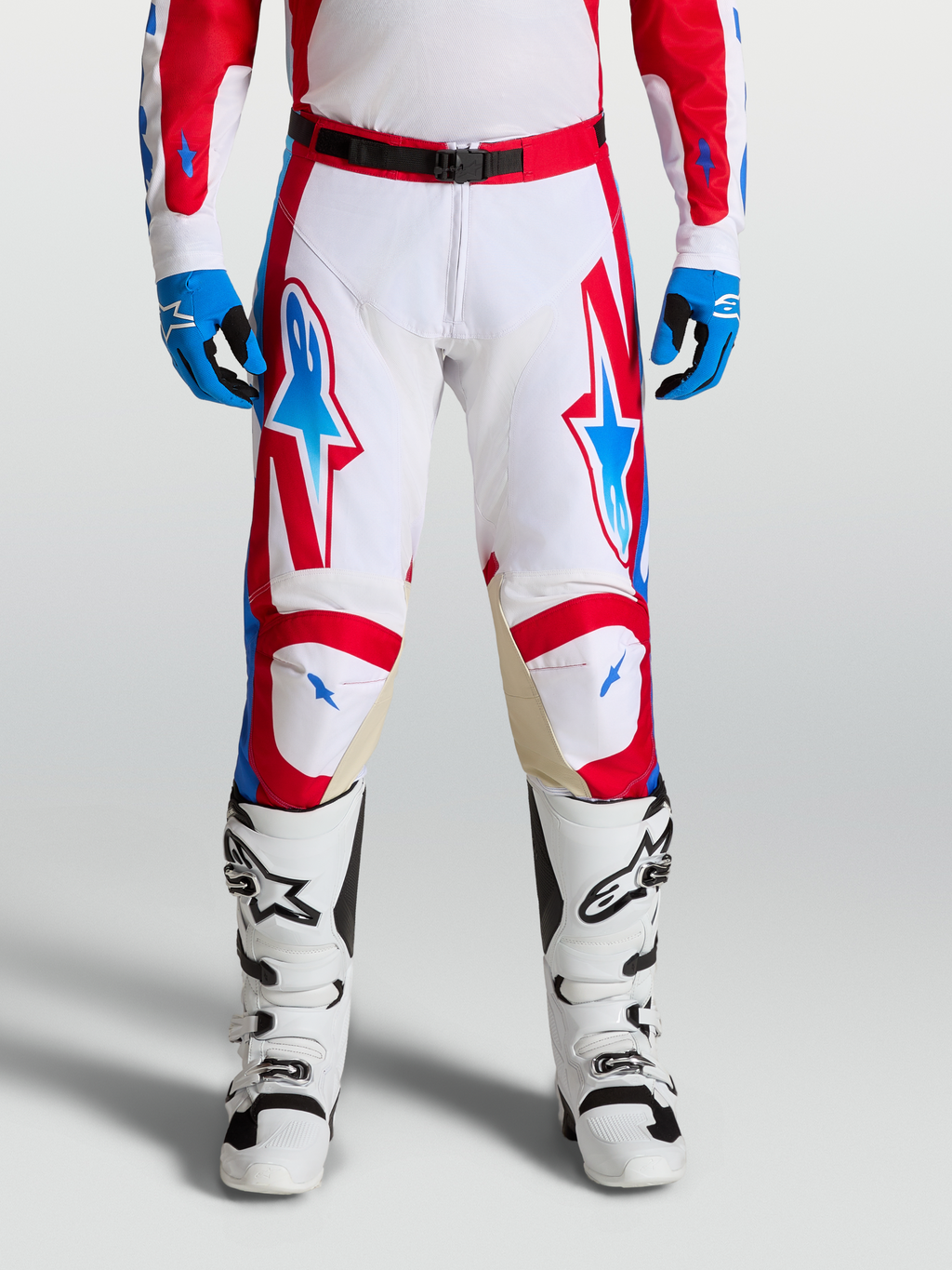 Alpinestars Pantaloni Racer Hunt, pantaloni motocross da uomo, bianco, rosso e blu, con gambe pre-curvate ergonomiche e pannelli in pelle sulle ginocchia per fuoristrada, indossati da un pilota con stivali bianchi.