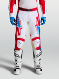 Alpinestars Pantaloni Racer Hunt, pantaloni motocross da uomo, bianco, rosso e blu, con gambe pre-curvate ergonomiche e pannelli in pelle sulle ginocchia per fuoristrada, indossati da un pilota con stivali bianchi.