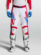 Alpinestars Pantaloni Racer Hunt, pantaloni motocross da uomo, bianco, rosso e blu, con gambe pre-curvate ergonomiche e pannelli in pelle sulle ginocchia per fuoristrada, indossati da un pilota con stivali bianchi.