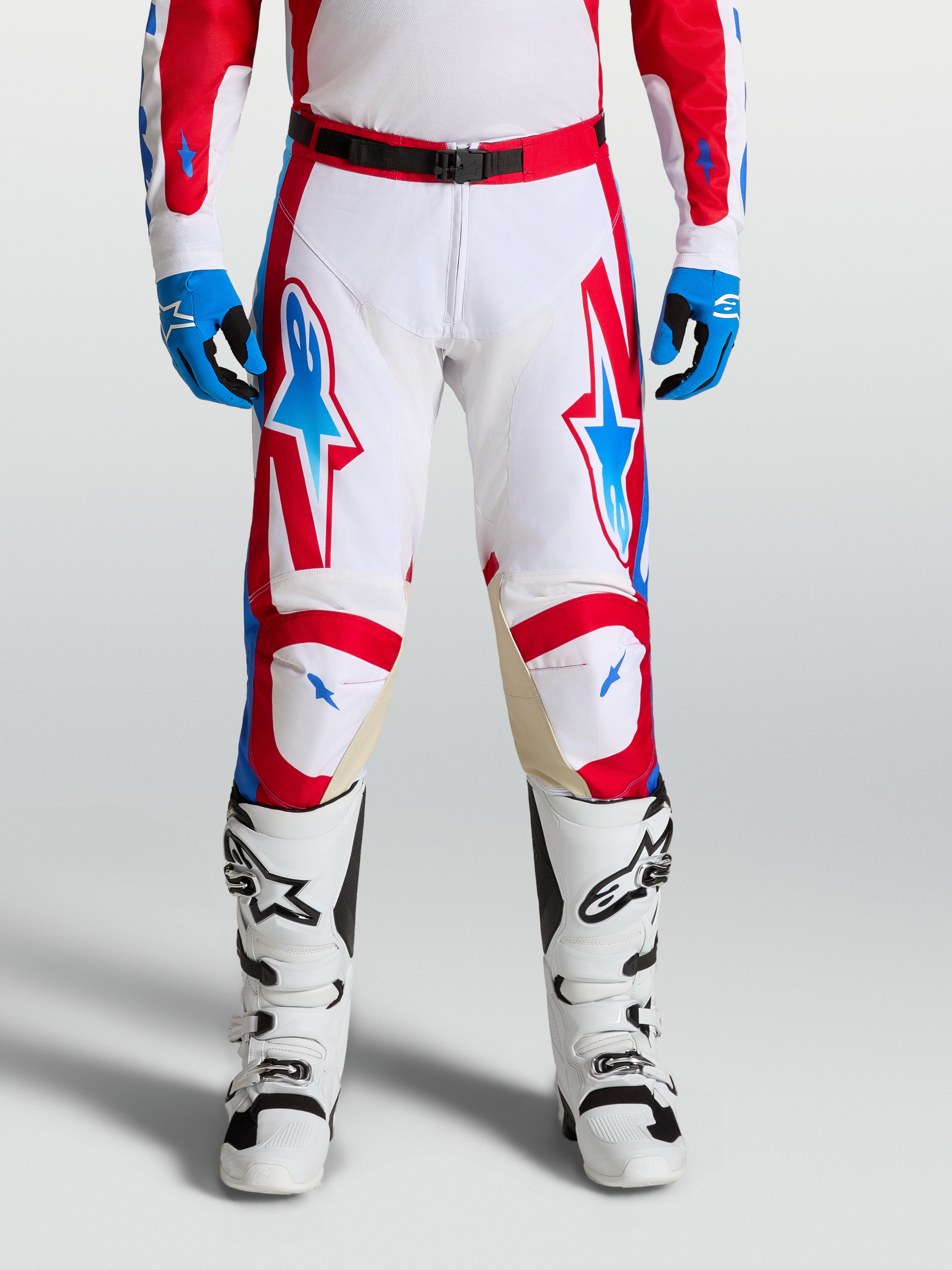 Alpinestars Pantaloni Racer Hunt, pantaloni motocross da uomo, bianco, rosso e blu, con gambe pre-curvate ergonomiche e pannelli in pelle sulle ginocchia per fuoristrada, indossati da un pilota con stivali bianchi.