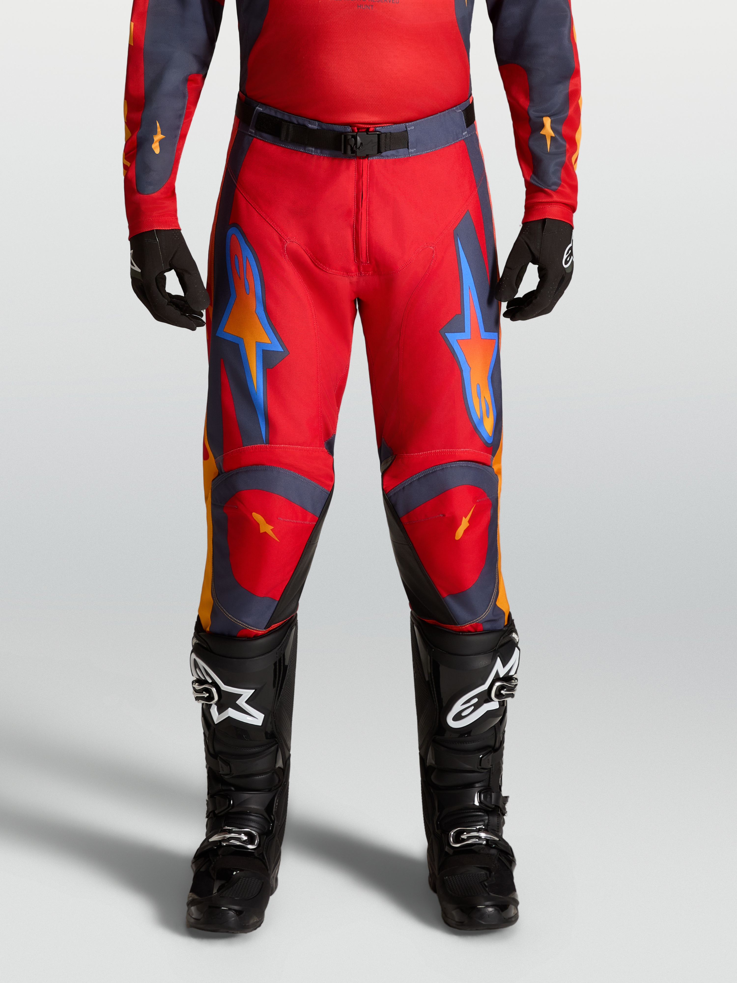 Alpinestars Pantaloni Racer Hunt, pantaloni motocross da uomo, rosso fuoco e grigio, vestibilità pre-curvata con ginocchia rinforzate, ideali per fuoristrada, indossati da un pilota con stivali neri.