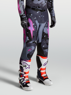 Alpinestars Pantaloni Racer Lone, pantaloni motocross da uomo, grigio camo e viola, design ergonomico precurvato e rinforzi in poliestere 600D, indossati con stivali Tech 10 bianchi e viola.
