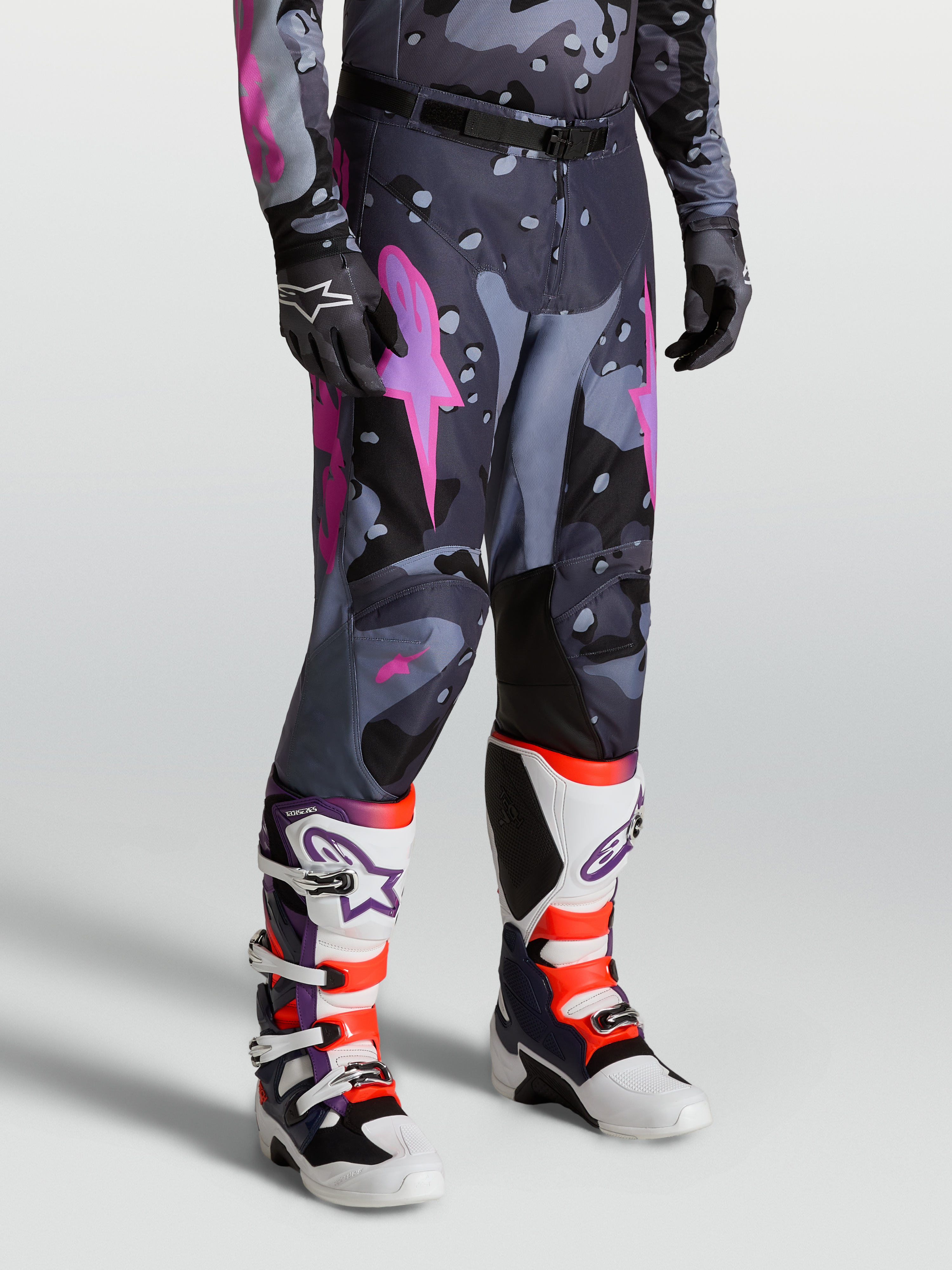 Alpinestars Pantaloni Racer Lone, pantaloni motocross da uomo, grigio camo e viola, design ergonomico precurvato e rinforzi in poliestere 600D, indossati con stivali Tech 10 bianchi e viola.