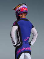 Pilota di motocross visto di spalle che indossa la maglia Techstar Knif 2026, nero, grigio chiaro e viola, con maniche bianche traforate e loghi Alpinestars rosa, abbinata a un casco coordinato viola e rosa, abbigliamento professionale da gara off-road.