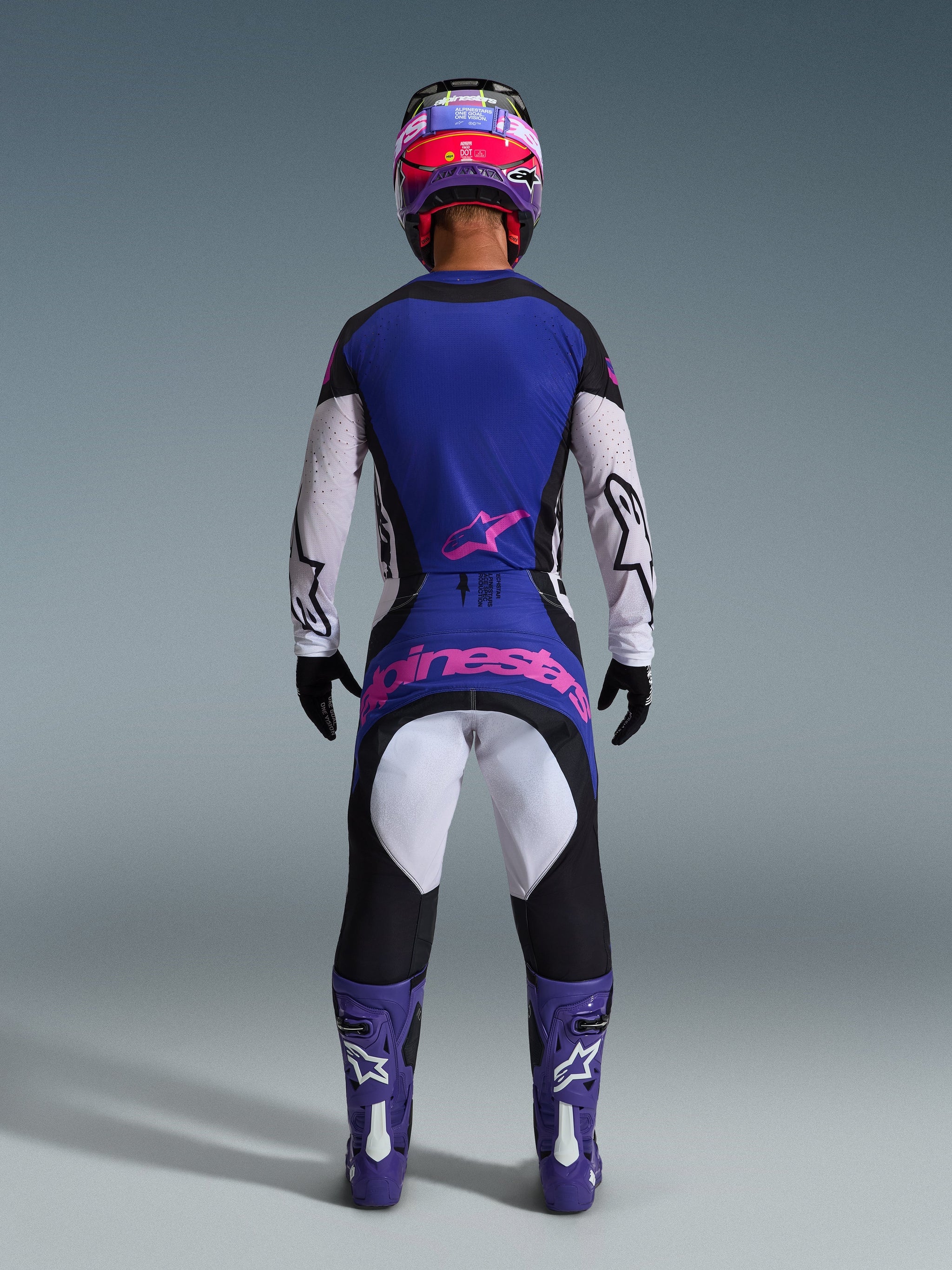 Pilota di motocross visto di spalle con maglia Techstar Knif 2026, nero grigio chiaro e viola, pantaloni da motocross coordinati con logo rosa, stivali e casco viola, abbigliamento off-road ad alte prestazioni