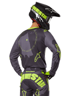 Pilota di motocross visto da dietro, completo Alpinestars 2025 Racer Hollow, maglia grigia con scritte circolari e loghi a stella giallo fluorescente, pantaloni grigi e gialli con branding evidente sulla seduta, casco nero e verde lime, abbigliamento per corse fuoristrada