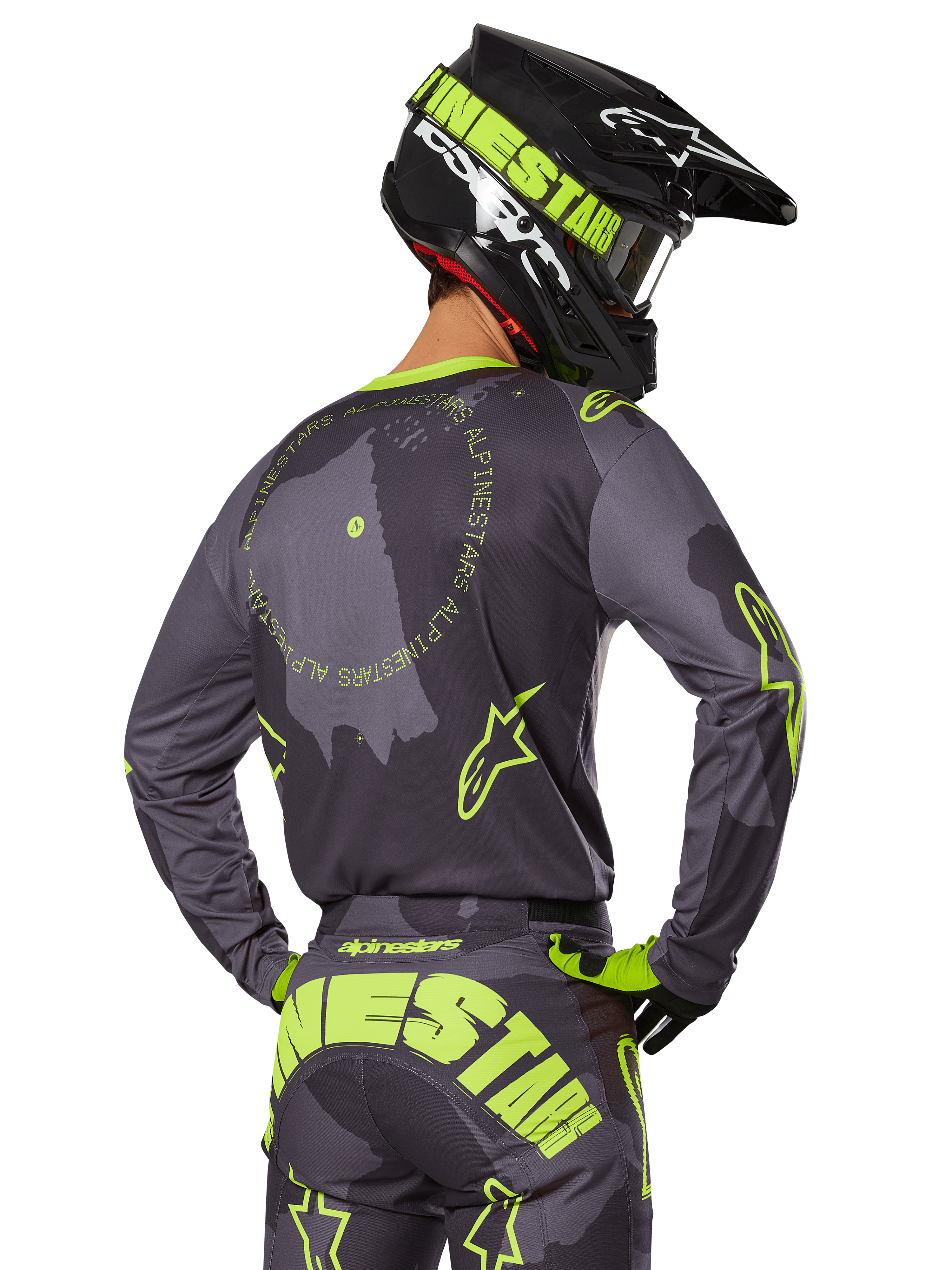 Pilota di motocross visto da dietro, completo Alpinestars 2025 Racer Hollow, maglia grigia con scritte circolari e loghi a stella giallo fluorescente, pantaloni grigi e gialli con branding evidente sulla seduta, casco nero e verde lime, abbigliamento per corse fuoristrada