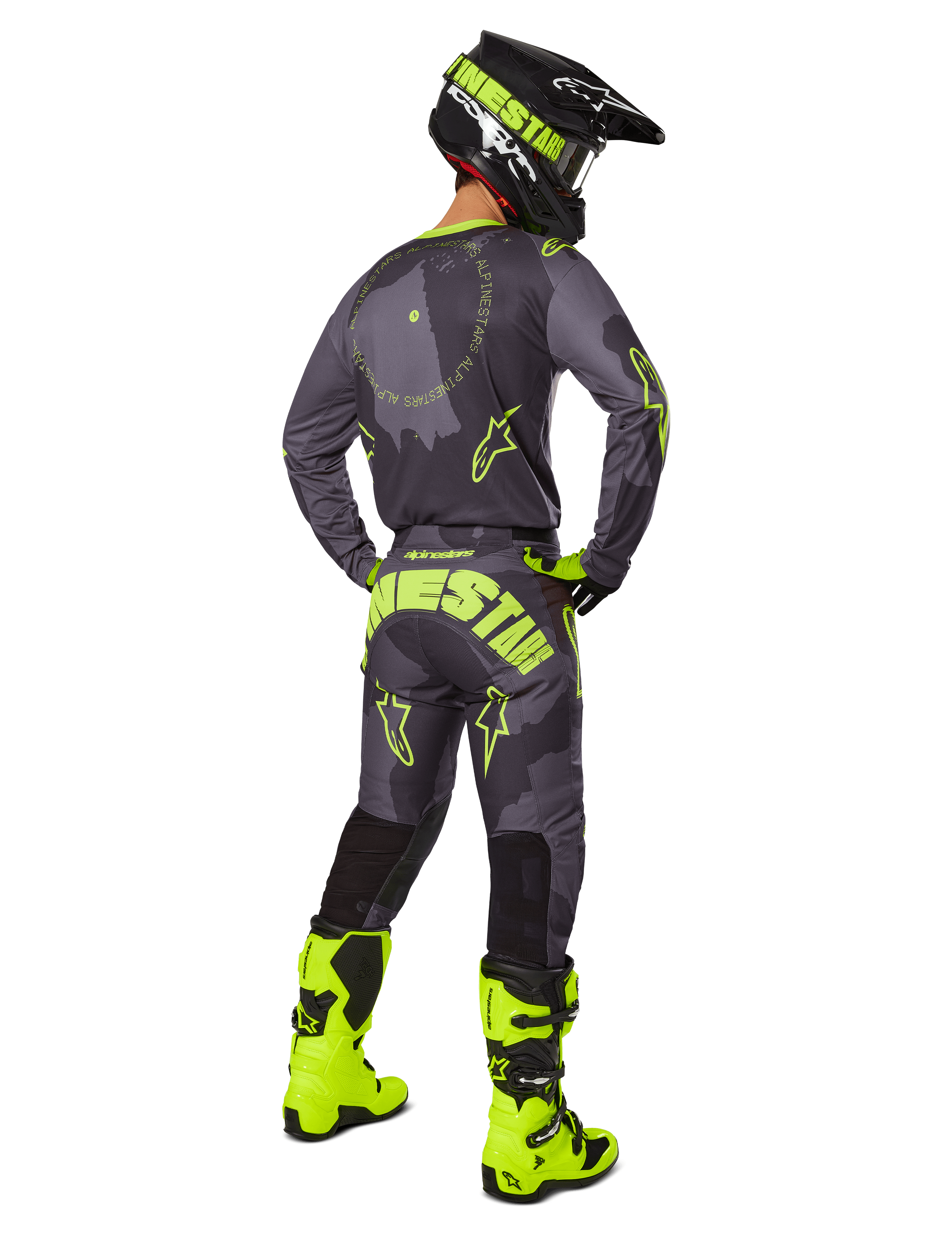 Vista posteriore di un pilota di motocross con il set completo Alpinestars Racer Hollow 2025 in Dark Camo Giallo Fluo, maglia camo grigia con dettagli giallo fluo, pantaloni coordinati con logo evidente sul retro, stivali giallo fluo e casco nero con maschera gialla.