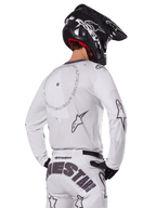 Pilota di motocross visto di spalle con il set completo Alpinestars 2025 Racer Hollow in bianco, maglia bianca a maniche lunghe con loghi a stella grigi e grafiche circolari, pantaloni bianchi coordinati con logo evidente sulla seduta, casco nero, abbigliamento da gara fuoristrada coordinato