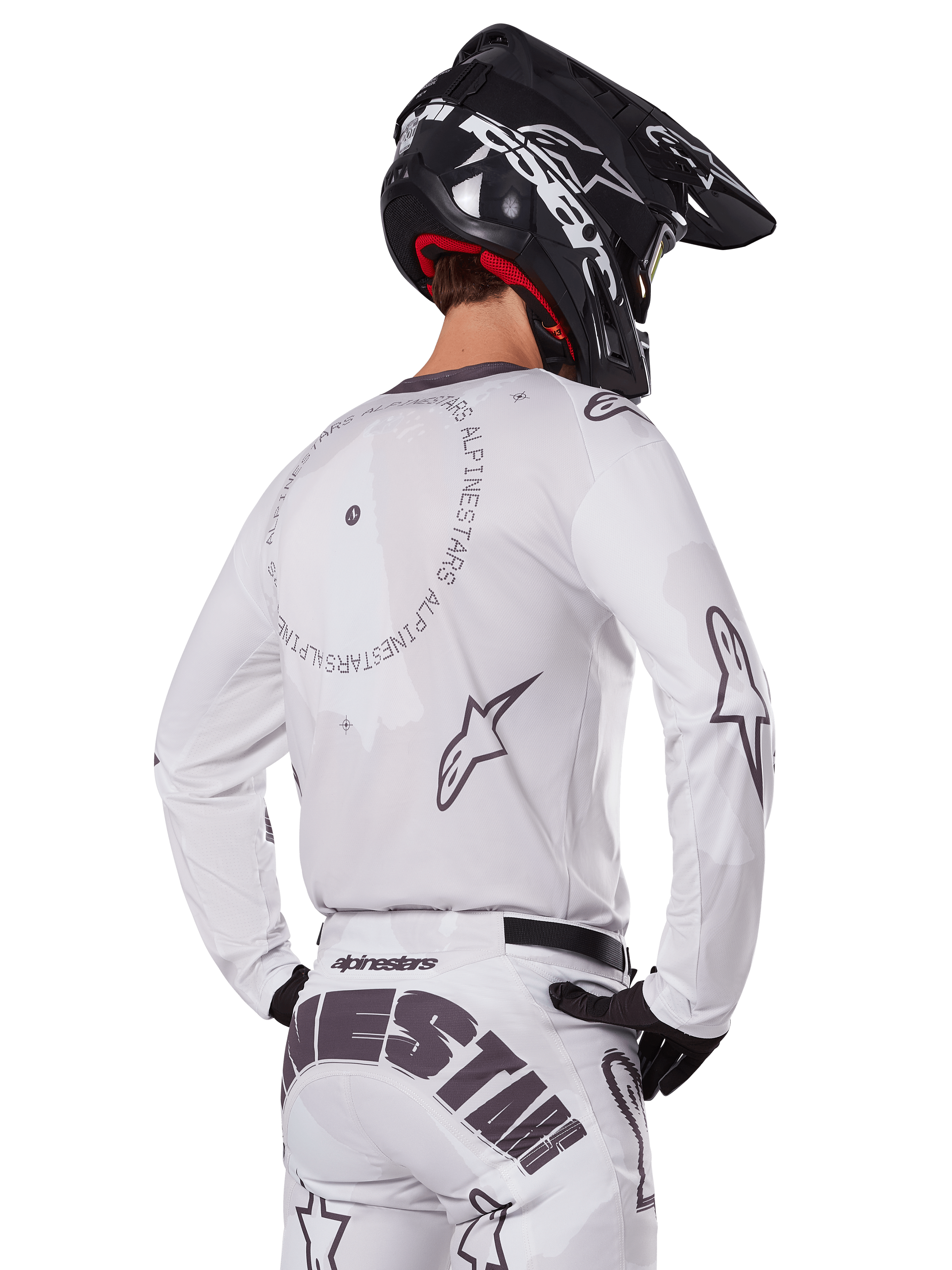Pilota di motocross visto di spalle con il set completo Alpinestars 2025 Racer Hollow in bianco, maglia bianca a maniche lunghe con loghi a stella grigi e grafiche circolari, pantaloni bianchi coordinati con logo evidente sulla seduta, casco nero, abbigliamento da gara fuoristrada coordinato