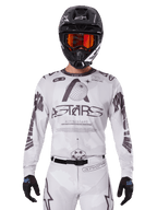 Alpinestars 2025 Racer Hollow Jersey, maglia da motocross bianca e nera, indossata con pantaloni MX bianchi coordinati e un casco nero con maschera arancione, caratterizzata da branding Alpinestars e grafiche a globo, abbigliamento professionale per corse fuoristrada
