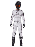 Set abbigliamento Alpinestars Racer Hollow 2025, bianco con grafiche nere e grigie, completo da motocross comprensivo di maglia e pantaloni, indossato con casco nero, occhiali arancioni, guanti neri e stivali neri, abbigliamento fuoristrada ad alte prestazioni