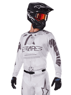 Alpinestars 2025 Racer Hollow Gear Set, maglia e pantaloni da motocross, bianco con grafiche nere e grigie, con marchio 'ASTARS' e motivi a globo, indossato con casco nero, occhiali a specchio rossi e guanti neri, progettato per prestazioni fuoristrada