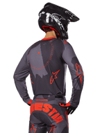 Maglia Alpinestars Racer Hollow 2025, vista posteriore di un pilota di motocross in completo Grigio Camo e Arancione Fluo, pantaloni coordinati con grande scritta Alpinestars arancione e casco nero, design professionale per fuoristrada