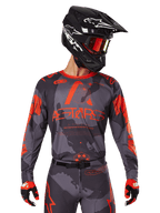 Alpinestars Maglia Racer Hollow 2025, maglia motocross grigio camo e arancione fluo, indossata con set coordinato di pantaloni e guanti, con casco nero con grafiche bianche e occhiali, branding ASTARS ben visibile, abbigliamento sportivo protettivo per corse fuoristrada
