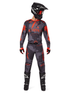 Pilota di motocross con maglia Alpinestars Racer Hollow 2025 e pantaloni abbinati in Grigio Camo Arancione Fluo, set completo con casco nero, occhiali, guanti e stivali, loghi arancioni e grafica mimetica