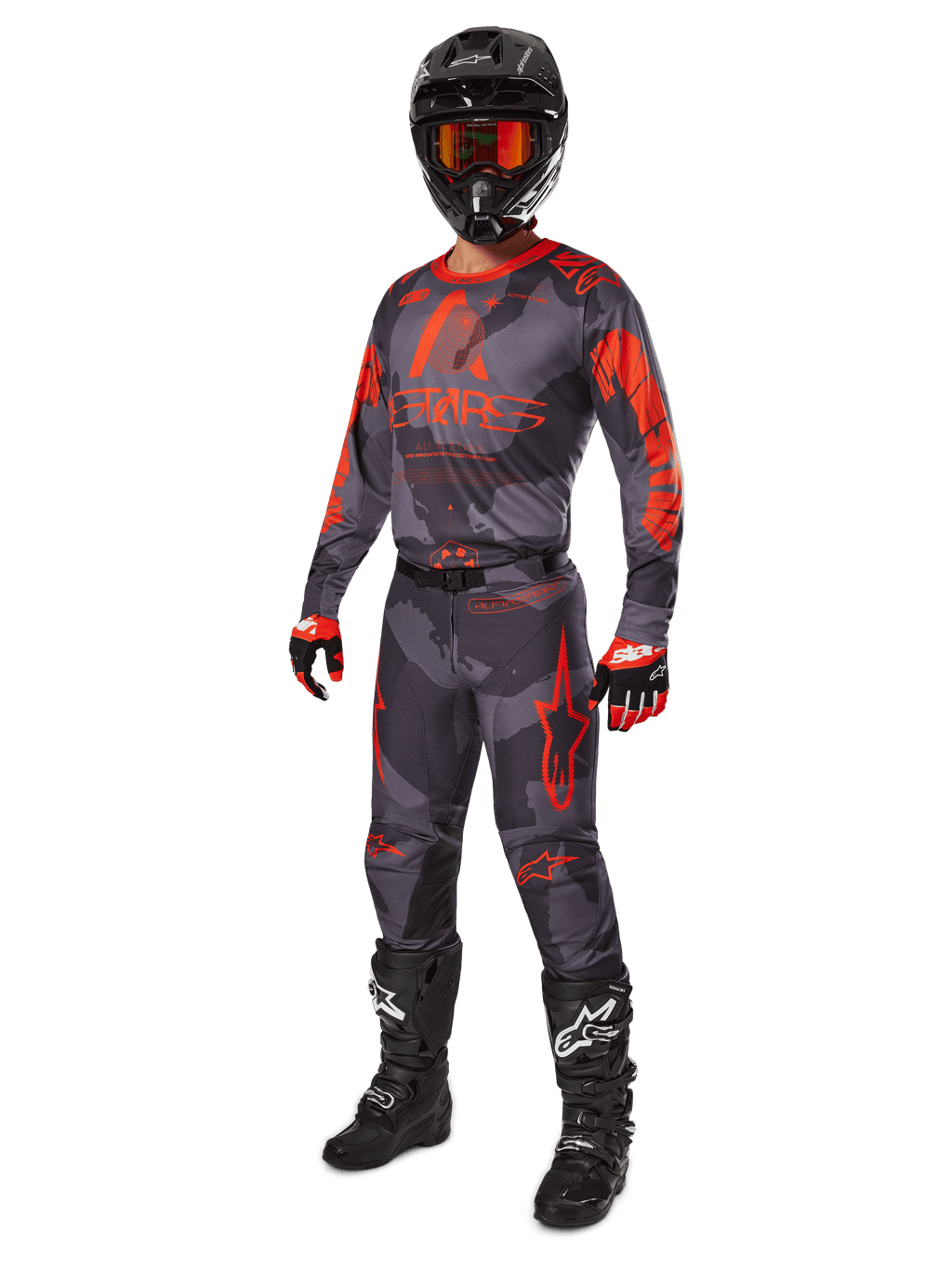 Pilota di motocross con abbigliamento da gara completo, maglia e pantaloni Alpinestars Racer Hollow 2025 in grigio camo arancione fluo, casco nero con occhiali arancioni, guanti neri e rossi e stivali neri con loghi bianchi, outfit coordinato per prestazioni fuoristrada