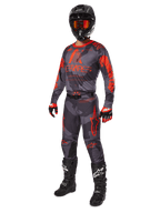 Pilota di motocross con abbigliamento da gara completo, maglia e pantaloni Alpinestars Racer Hollow 2025 in grigio camo arancione fluo, casco nero con occhiali arancioni, guanti neri e rossi e stivali neri con loghi bianchi, outfit coordinato per prestazioni fuoristrada