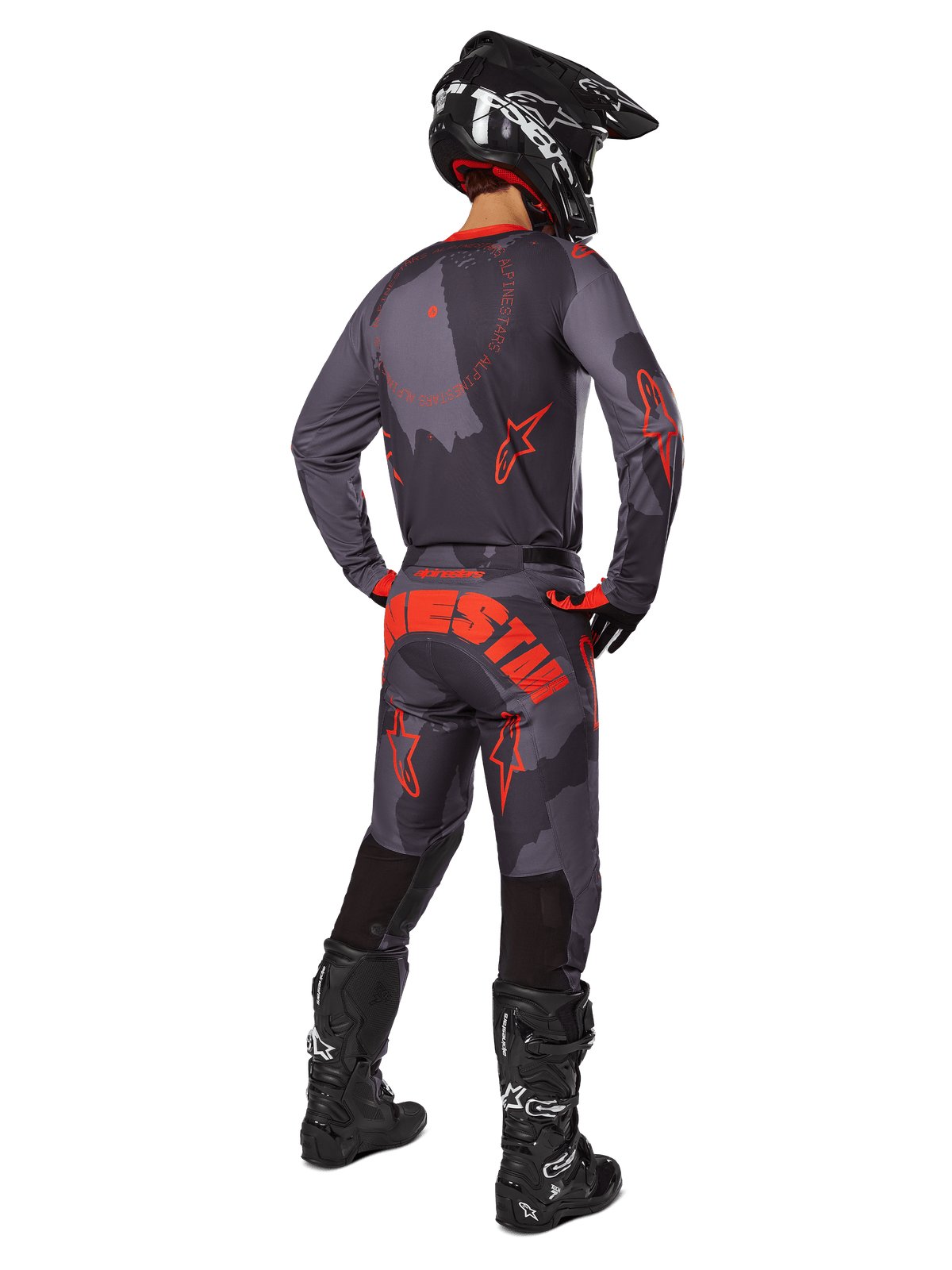 Pilota di motocross visto da dietro che indossa la maglia Alpinestars Racer Hollow 2025 in Gray Camo Orange Fluo, pantaloni mimetici coordinati con scritta Alpinestars arancione, casco nero e stivali neri, completo coordinato per la guida off-road