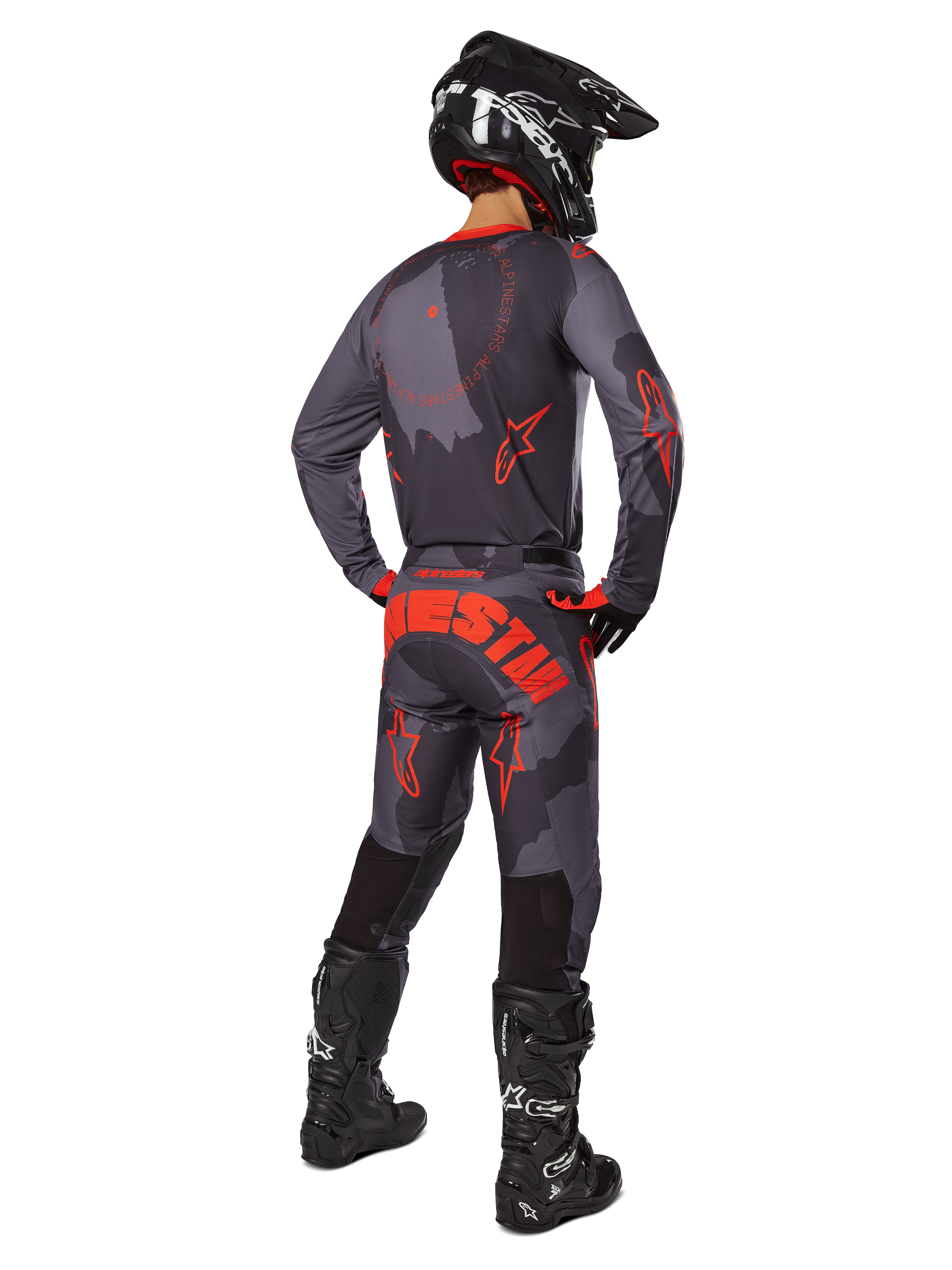 Pilota di motocross visto da dietro che indossa la maglia Alpinestars Racer Hollow 2025 in Gray Camo Orange Fluo, pantaloni mimetici coordinati con scritta Alpinestars arancione, casco nero e stivali neri, completo coordinato per la guida off-road