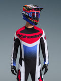 Alpinestars 2026 Techstar Knif Jersey, maglia da motocross, rosso, bianco e blu, con un vistoso logo sul petto e busto sfumato, indossata da un pilota con casco multicolore coordinato, occhiali blu e guanti neri, abbigliamento professionale da fuoristrada.