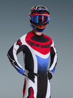 Pilota di motocross che indossa la maglia Alpinestars Techstar Knif 2026 in rosso, bianco e blu, con marchio nero sul petto, abbinata a pantaloni da motocross coordinati, casco da corsa con occhiali blu e guanti neri e bianchi, attrezzatura professionale da fuoristrada per competizioni