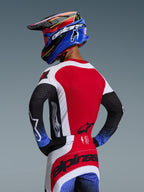 Pilota di motocross visto da dietro con il set completo Techstar Knif 2026, maglia rossa con pannelli laterali bianchi e maniche nere con polsini blu puntinati, pantaloni rossi e bianchi con logo Alpinestars nero sulla seduta, casco coordinato blu e rosso con maschera.