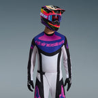 Pilota di motocross con completo Alpinestars Techstar Knif 2026, maglia con grafica blu e rosa sul petto e busto bianco, pantaloni bianchi con pannelli neri e loghi rosa, casco multicolore viola e rosa con occhiali coordinati, guanti neri, abbigliamento professionale per gare fuoristrada