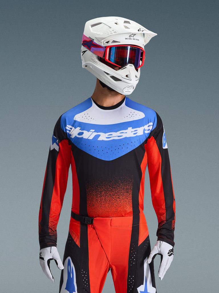 Alpinestars Techstar Knif Jersey 2026, maglia da motocross, arancione, nero e blu UCLA, con pannello blu sul petto, logo Alpinestars bianco e maniche arancioni-rosse, indossata con casco bianco, occhiali rossi e guanti bianchi, abbigliamento professionale da fuoristrada