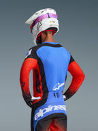 Pilota di motocross visto da dietro che indossa il completo Techstar Knif 2026, maglia blu con maniche rosse e accenti neri, pantaloni neri e blu con marchio Alpinestars bianco, casco bianco con elastico degli occhiali rosa, con traforature al laser per la ventilazione