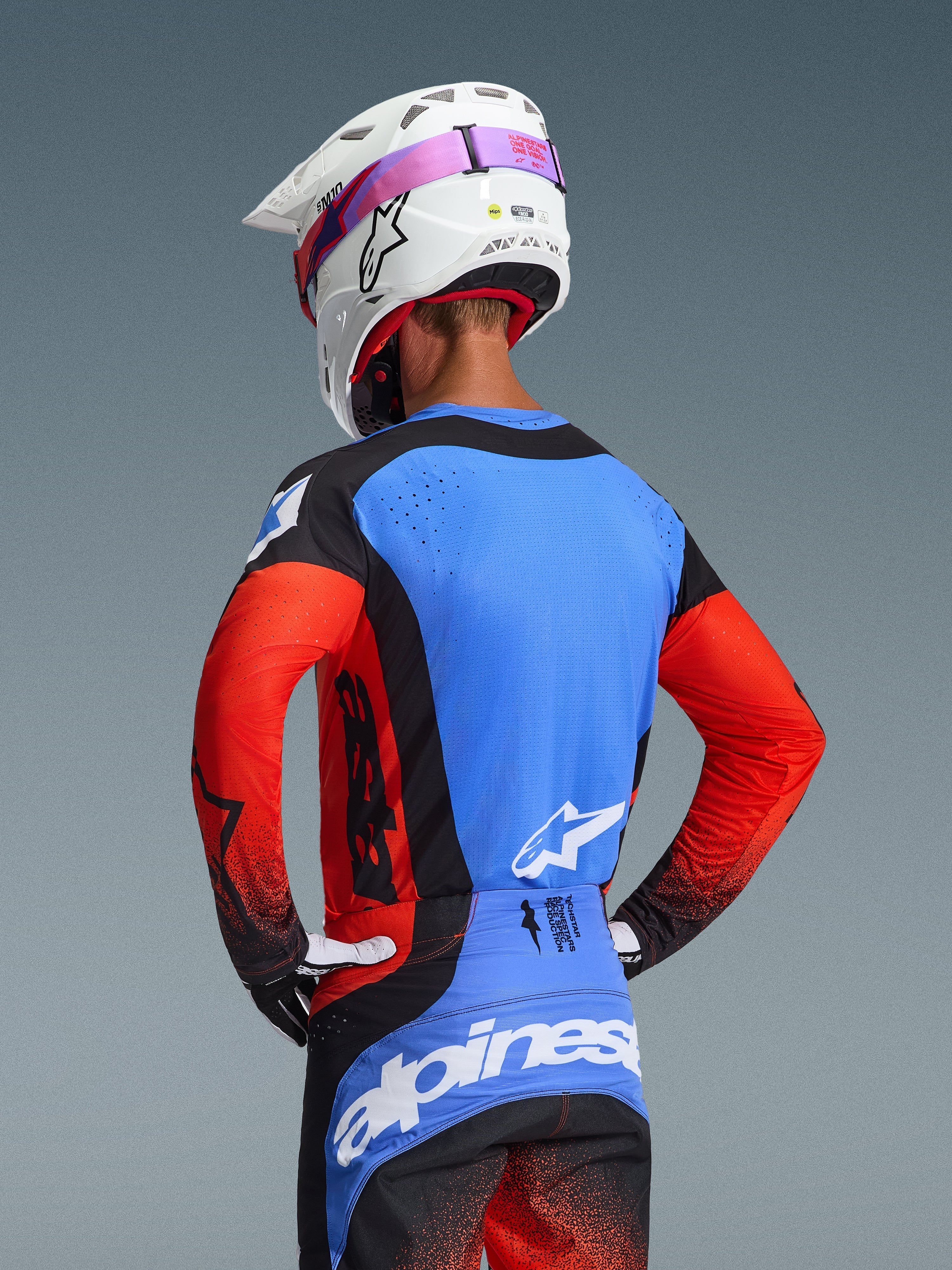 Vista posteriore di un pilota di motocross che indossa la maglia Techstar Knif 2026 in Orange Black UCLA Blue, con pannello posteriore blu e perforazioni tagliate al laser, maniche arancioni e dettagli neri, abbinata a pantaloni da motocross coordinati e casco Alpinestars bianco.
