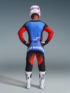 Vista posteriore di un pilota di motocross con il completo Alpinestars 2026 Techstar Knif Gear Set, maglia blu e rossa con dettagli neri, pantaloni neri con sfumatura rossa sulle cosce e logo blu, abbinato a stivali da corsa bianchi e casco bianco con maschera rosa.