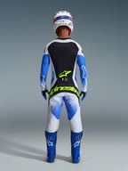 Alpinestars 2026 Techstar Knif Gear Set, vista posteriore di un pilota di motocross in blu Ucla, nero e bianco, con branding giallo fluo su maglia e pantaloni, abbinato a stivali da gara blu e casco bianco.