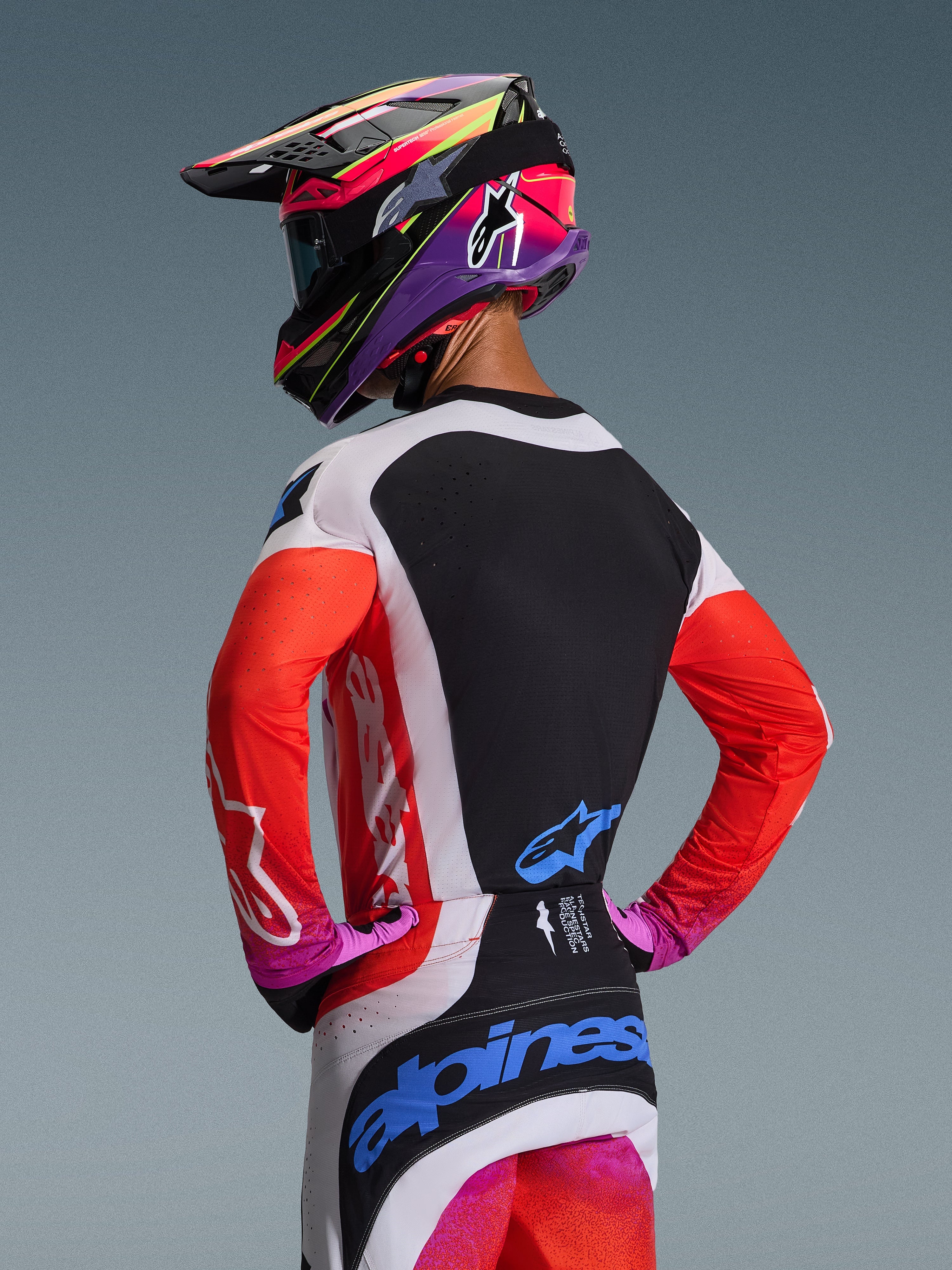 Pilota di motocross visto di spalle con completo Alpinestars Techstar Knif 2026, maniche arancione fluo e pannello posteriore nero, pantaloni bianchi e neri con loghi blu, casco viola e rosa vivace, abbigliamento professionale per prestazioni off-road