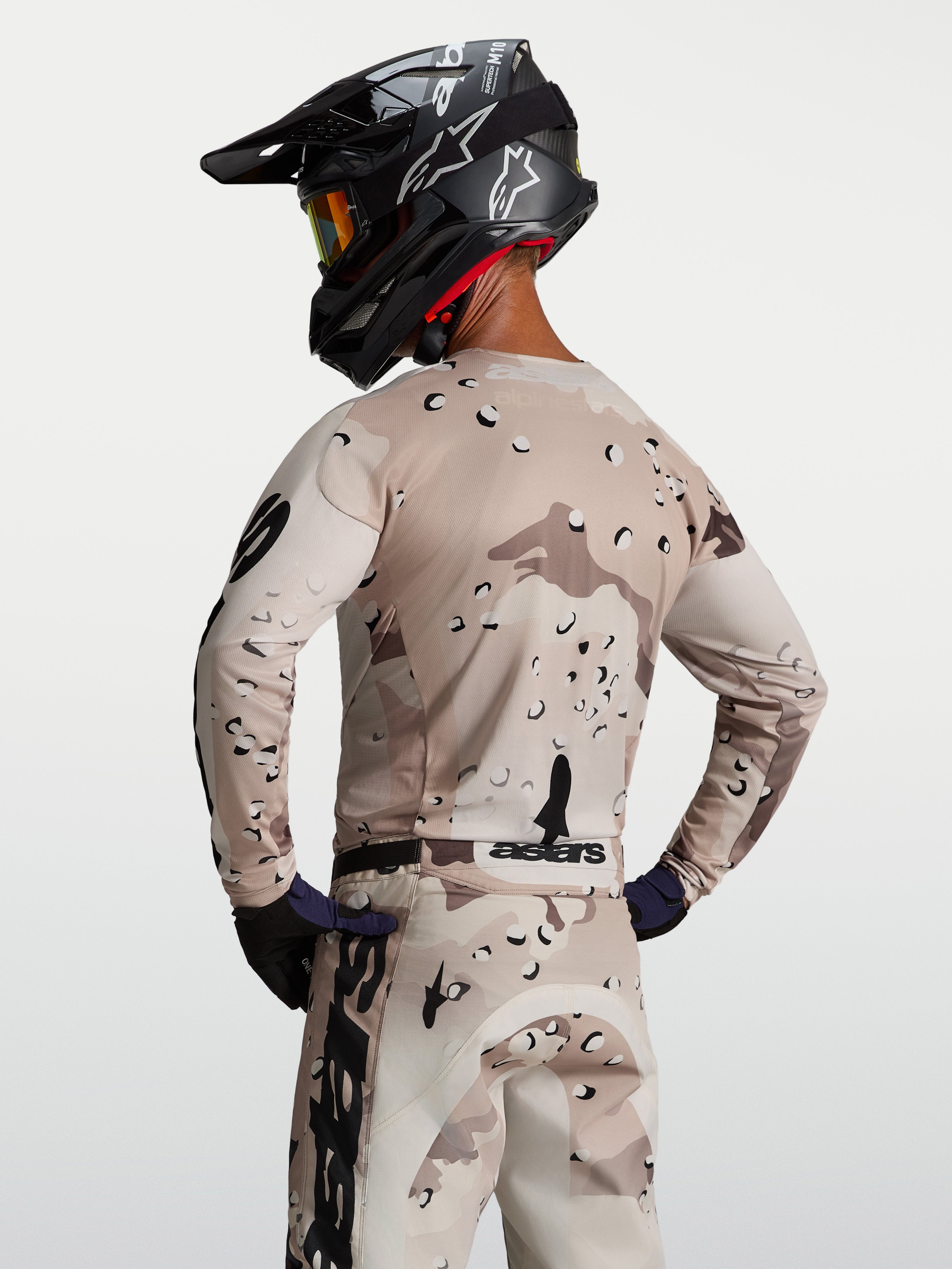 Alpinestars Maglia Racer Lone, maglia da motocross da uomo, sabbia e nero con motivo desert camo, vista posteriore su modello con casco, tessuto tecnico traspirante per sessioni off-road e enduro