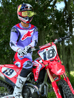 Pilota di motocross su una moto Honda CRF rossa, indossa la maglia Alpinestars 2025 Fluid Wurx in viola e rosso acceso, pantaloni coordinati viola e rossi, casco Red Bull con occhiali gialli, stivali e guanti Alpinestars bianchi, equipaggiamento professionale da MX per guida fuoristrada