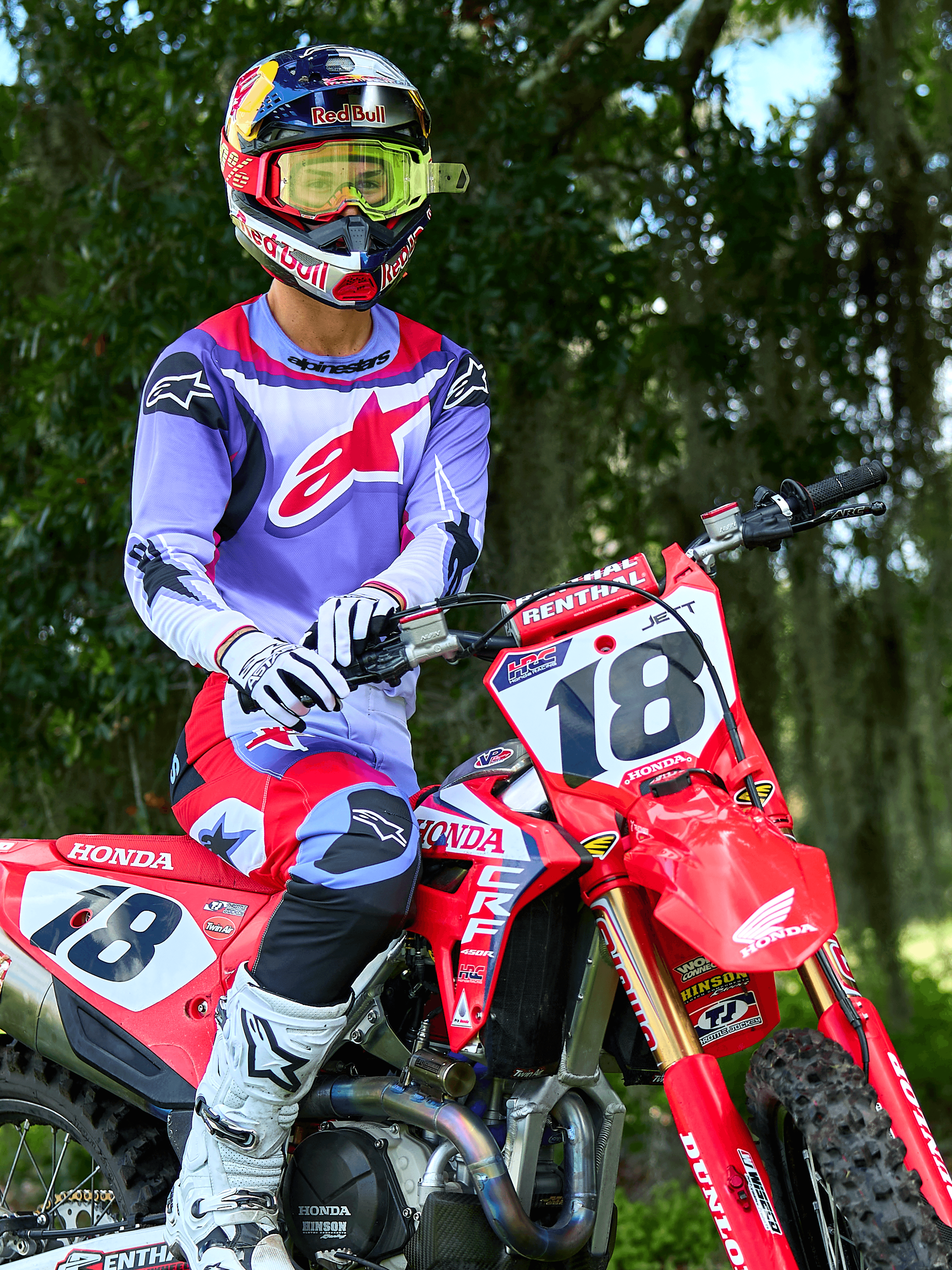 Pilota di motocross su una moto Honda CRF rossa, indossa la maglia Alpinestars 2025 Fluid Wurx in viola e rosso acceso, pantaloni coordinati viola e rossi, casco Red Bull con occhiali gialli, stivali e guanti Alpinestars bianchi, equipaggiamento professionale da MX per guida fuoristrada