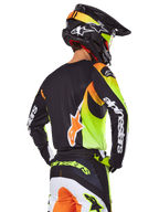 Pilota di motocross visto da dietro con set Alpinestars 2025 Fluid Wurx, maglia nera con grafiche giallo neon e arancione, pantaloni coordinati con pannelli bianchi e arancioni, casco nero con accenti neon, branding Alpinestars su maniche e seduta