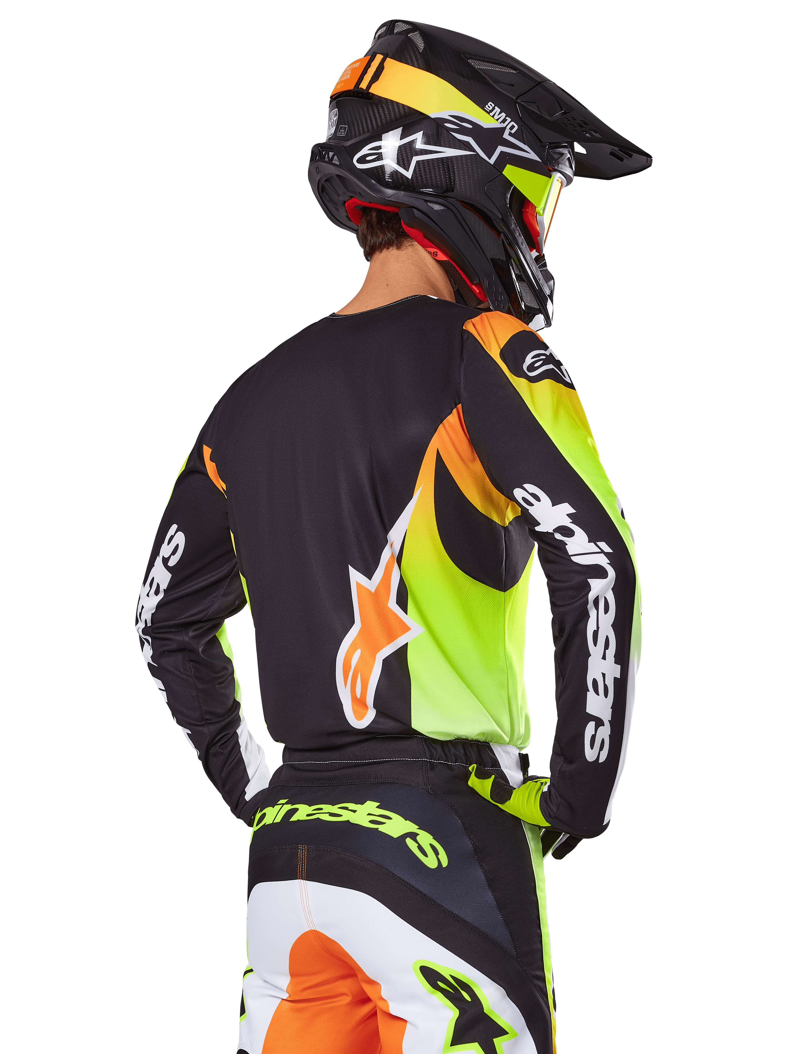 Pilota di motocross visto da dietro con set Alpinestars 2025 Fluid Wurx, maglia nera con grafiche giallo neon e arancione, pantaloni coordinati con pannelli bianchi e arancioni, casco nero con accenti neon, branding Alpinestars su maniche e seduta