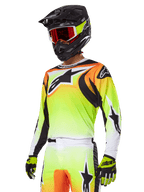 Pilota di motocross con completo Alpinestars 2025 Fluid Wurx, maglia con sfumatura gialla e arancione con dettagli neri e grandi loghi a stella, pantaloni da motocross coordinati, casco integrale nero con maschera a lenti rosse e guanti gialli, outfit ad alte prestazioni per competizioni off-road