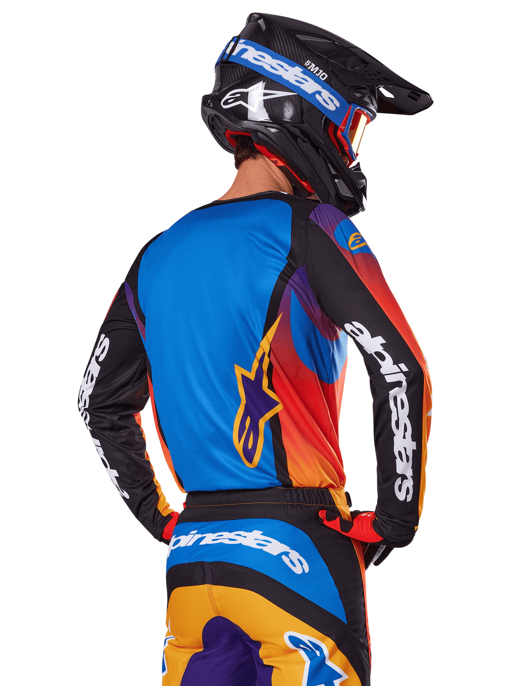 Pilota di motocross visto dal retro con indosso il set Alpinestars 2025 Fluid Wurx, maglia multicolore con retro blu e maniche nere con loghi bianchi, pantaloni arancioni e blu coordinati, casco nero e blu con occhiali arancioni, completo da gara off-road.