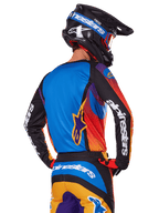 Pilota di motocross visto dal retro con indosso il set Alpinestars 2025 Fluid Wurx, maglia multicolore con retro blu e maniche nere con loghi bianchi, pantaloni arancioni e blu coordinati, casco nero e blu con occhiali arancioni, completo da gara off-road.