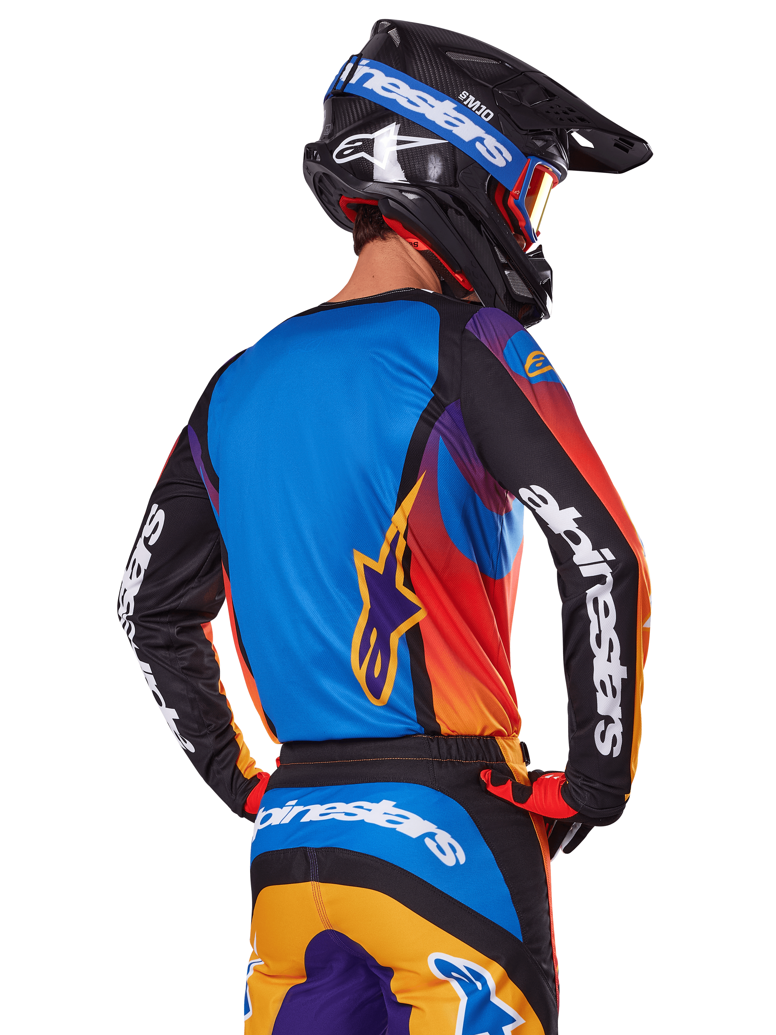 Pilota di motocross visto dal retro con indosso il set Alpinestars 2025 Fluid Wurx, maglia multicolore con retro blu e maniche nere con loghi bianchi, pantaloni arancioni e blu coordinati, casco nero e blu con occhiali arancioni, completo da gara off-road.
