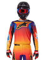 Alpinestars 2025 Fluid Wurx Jersey, maglia da motocross multicolore con sfumatura arancione e rossa e dettagli viola, indossata con equipaggiamento coordinato, casco nero con occhiali blu e rossi e guanti rossi, con un grande logo a stella nero sul petto.