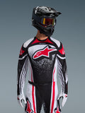 Alpinestars Maglia Techstar Nomur 2026, maglia da motocross, nero, bianco e rosso, indossata da un pilota con pantaloni coordinati, casco nero e guanti bianchi, con un grande logo Astars rosso e grafica punteggiata, vestibilità atletica da gara per competizioni off-road