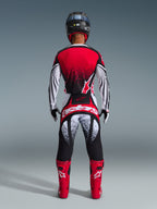 Pilota di motocross visto da dietro che indossa il completo Alpinestars 2026 Techstar Nomur, maglia rossa e bianca con sfumatura punteggiata nera, pantaloni coordinati bianchi e rossi con dettagli neri, stivali rosso acceso e casco nero, equipaggiamento da gara fuoristrada coordinato.