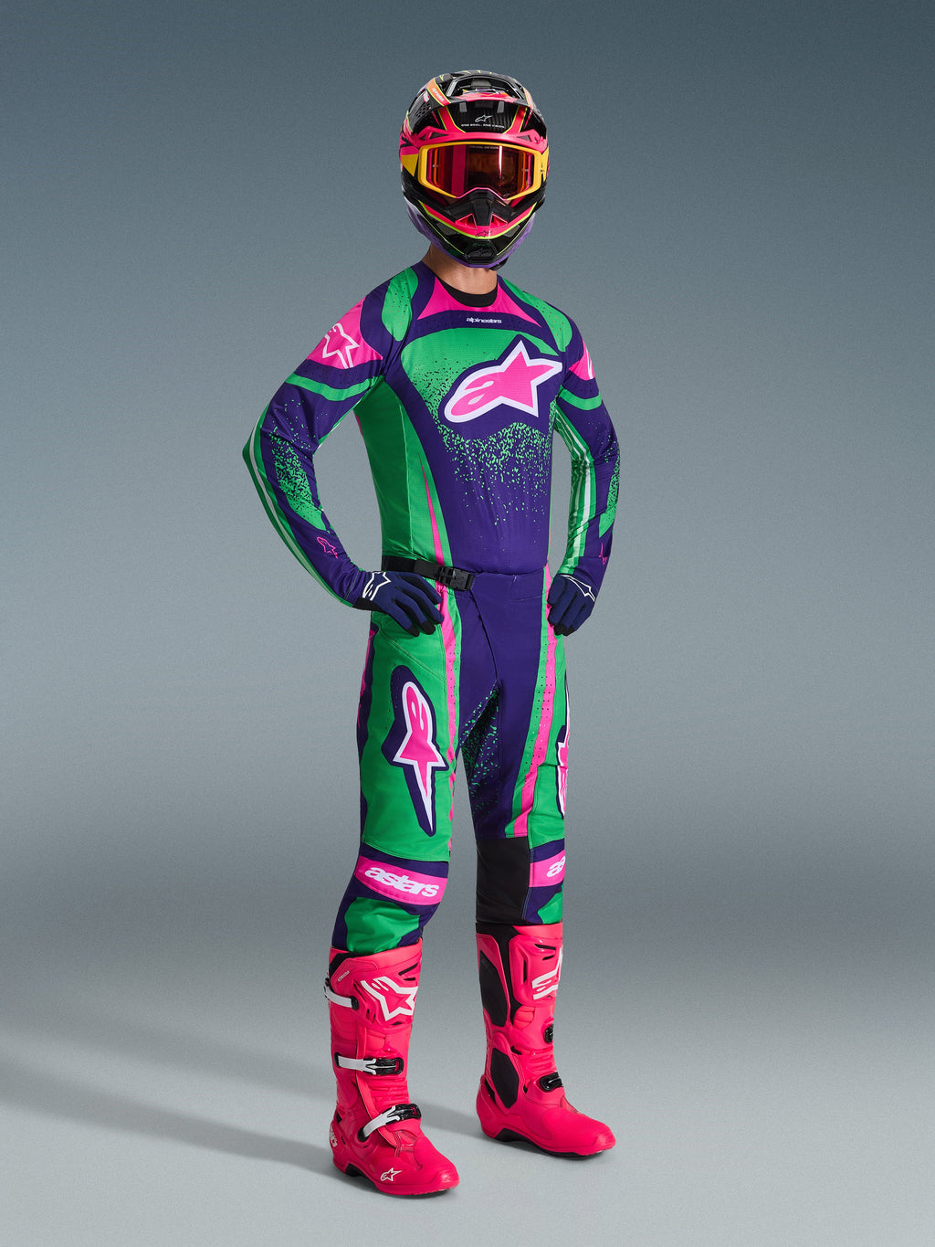 Pilota di motocross che indossa il set completo Alpinestars 2026 Techstar Nomur in viola scuro, verde e rosa fluo, composto da maglia e pantaloni coordinati, casco colorato con maschera rosa e vivaci stivali rosa, equipaggiamento professionale per corse fuoristrada.